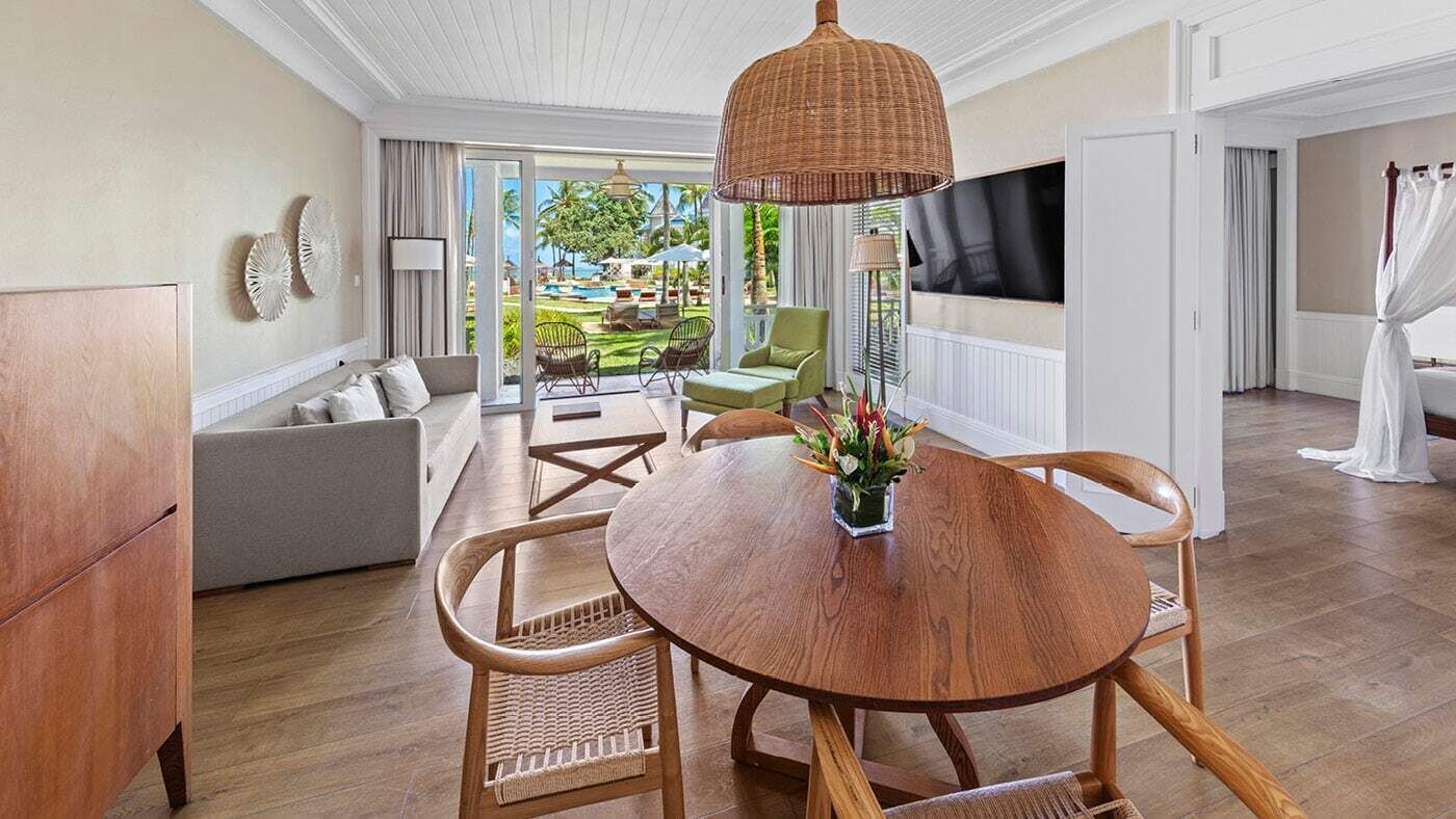 Una habitación con una mesa redonda de madera, sillas de mimbre, un sofá y un ventanal con vistas a una piscina y vegetación.