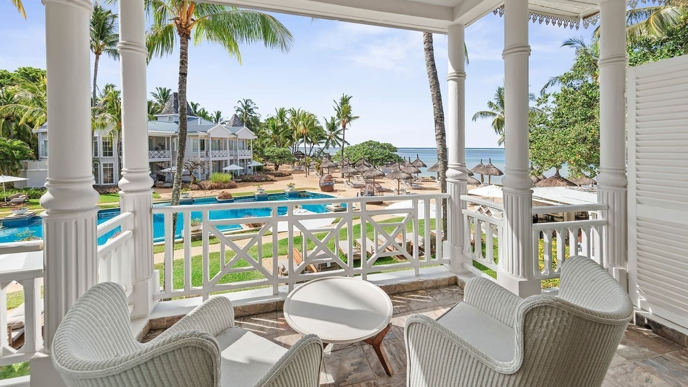 A covered outdoor area with a pool, white wicker furniture, and a view of a beach and ocean.