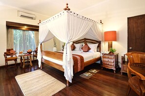 A four-poster canopy bed with a wooden frame and white drapes, a bedside table with a lamp, and a view of the outdoors through a window with curtains.