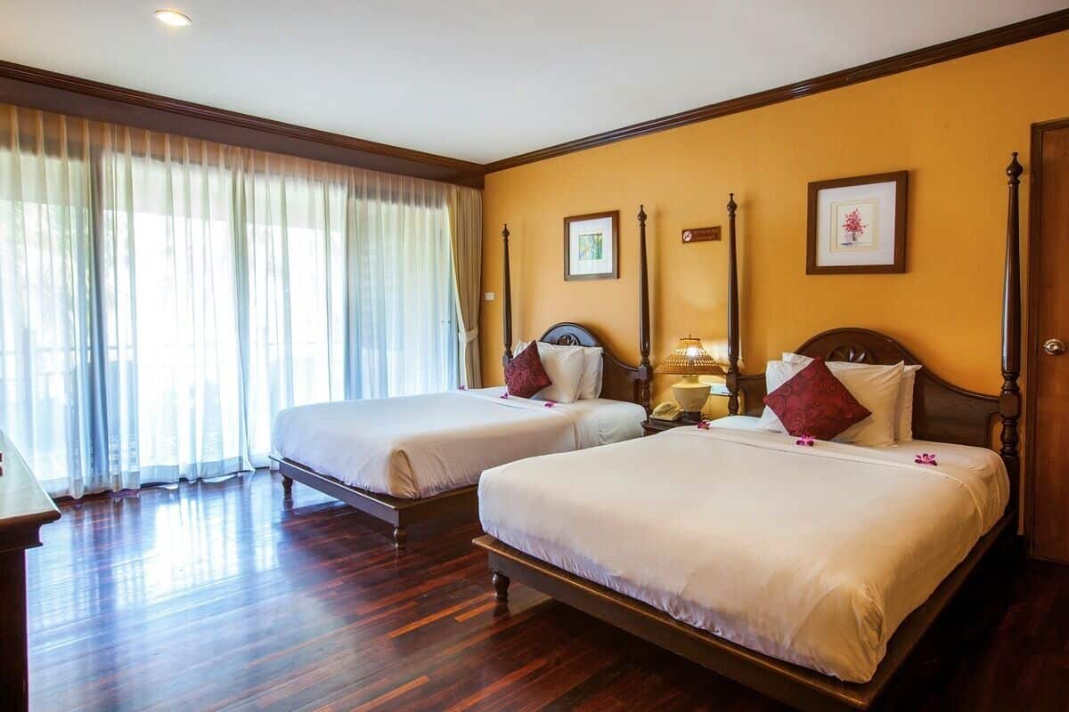 Een hotelkamer met twee bedden, houten vloeren, een groot raam met dunne gordijnen en kunstwerken in lijsten aan de muren.