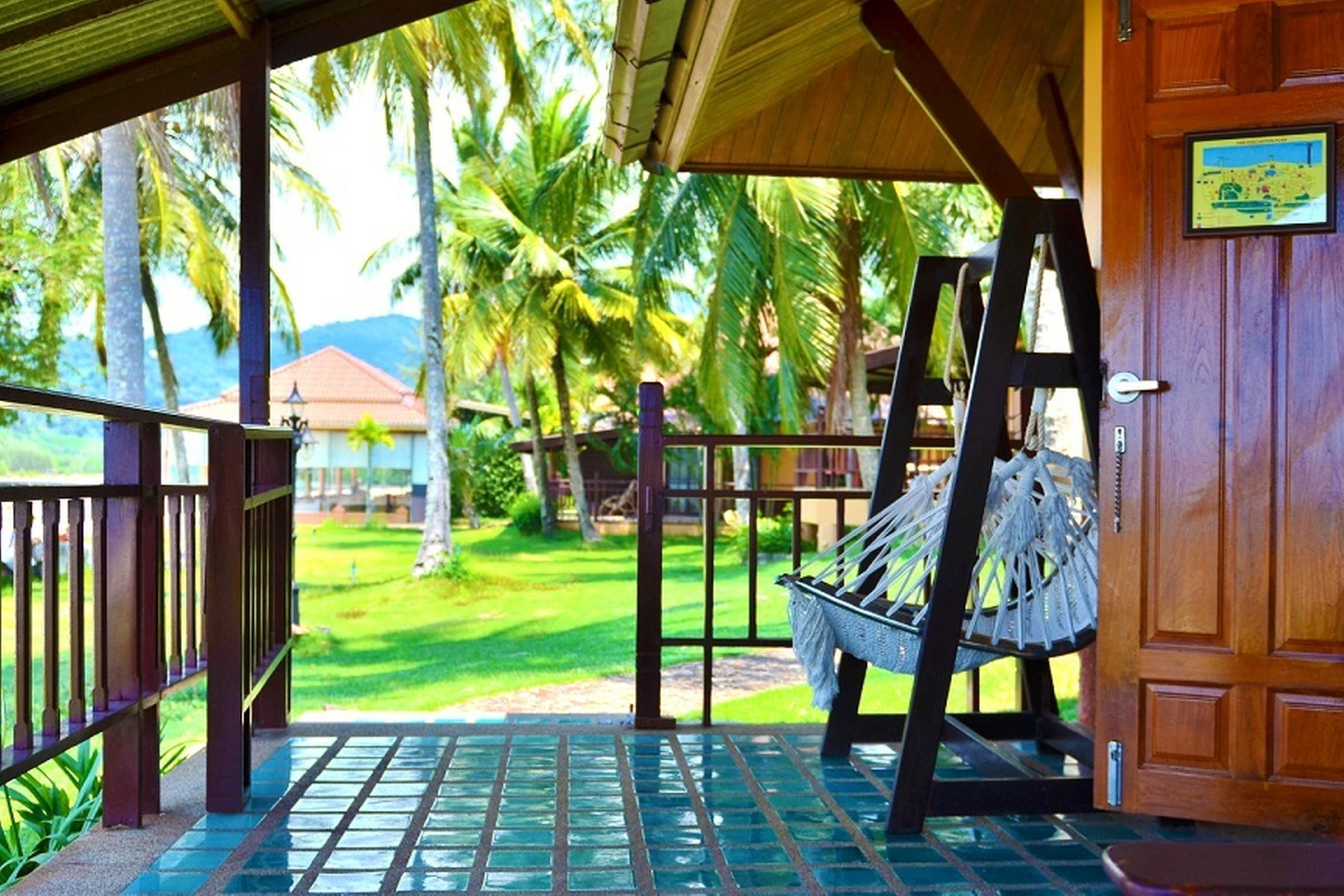A wooden porch with a hammock, a wooden door, and a view of a well-maintained lawn with palm trees.