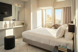 A hotel room with a bed, a small table with books, a chair, a mirror, and a window with a view of buildings.