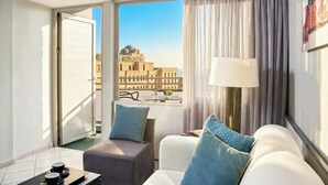 Ein Hotelzimmer mit Balkon, einer Couch mit blauen Kissen, einem Tisch mit Lampe und Blick auf ein Gebäude mit Kuppel.