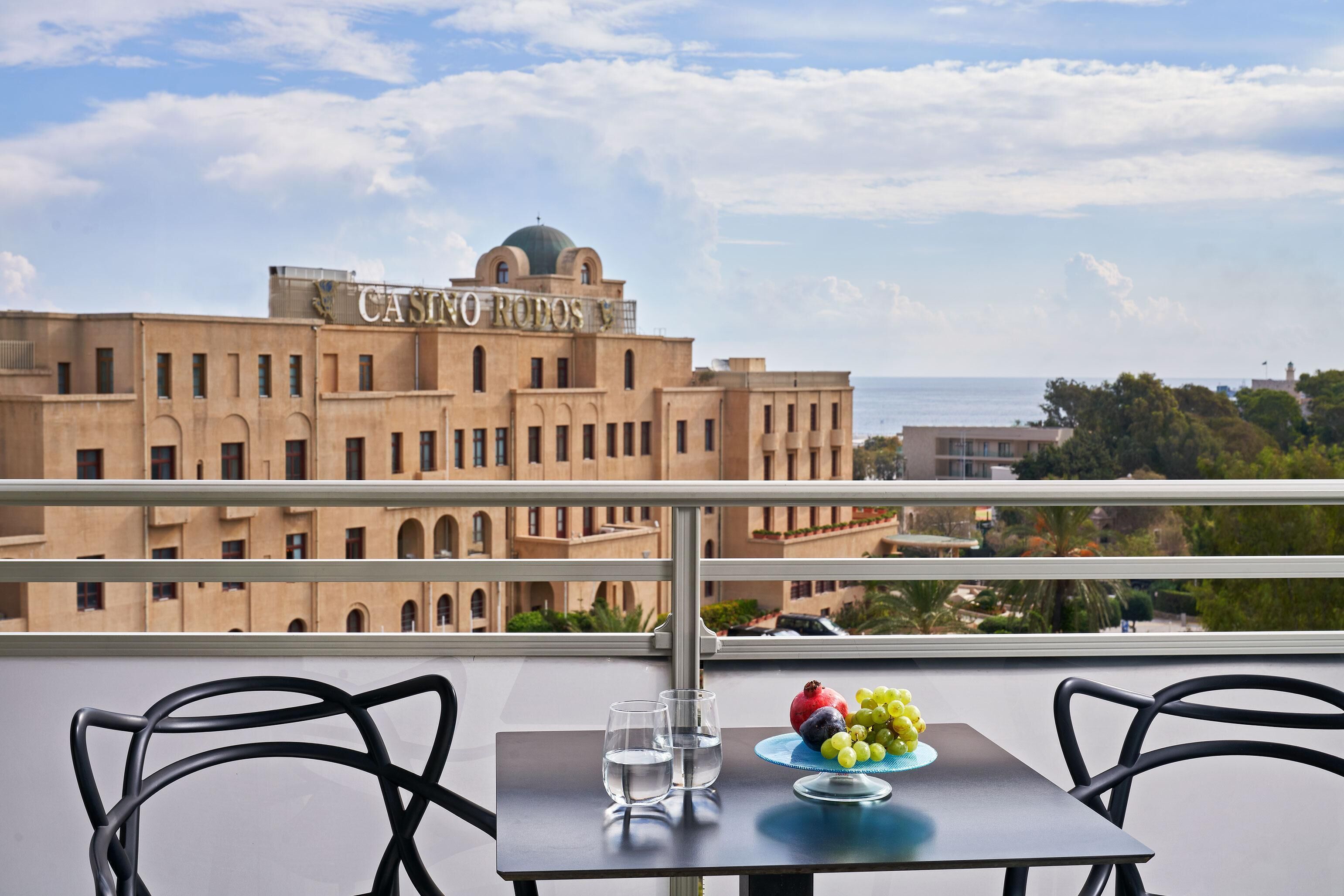 Un balcón con una mesa, dos sillas, un plato de uvas y una granada, y dos vasos de agua. Al fondo, un edificio con una cúpula y el letrero 'CASINO RODOS'.