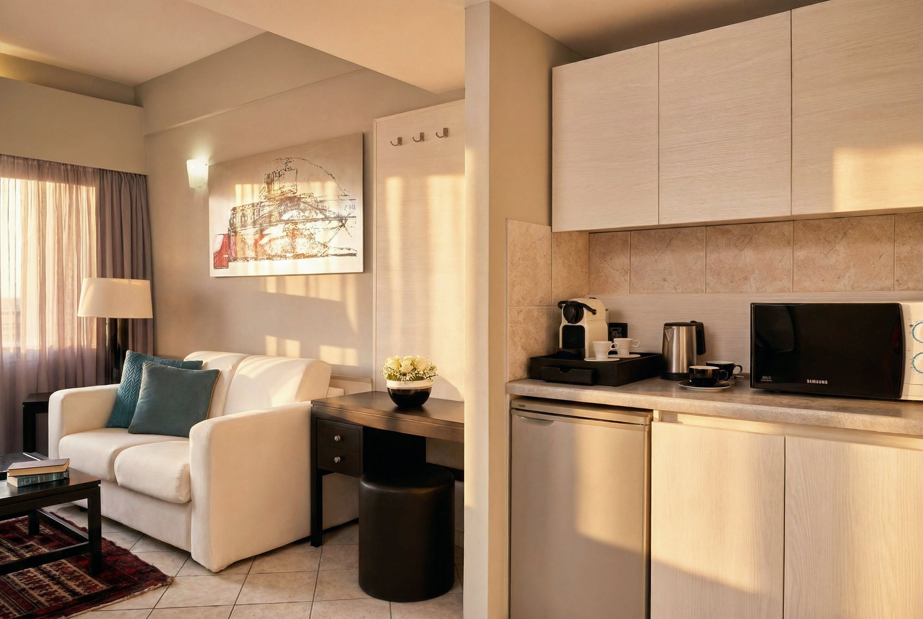 A modern living room with a sofa, coffee table, and a small side table. There is a kitchen area with a microwave, coffee maker, and cabinets. A painting is visible on the wall.