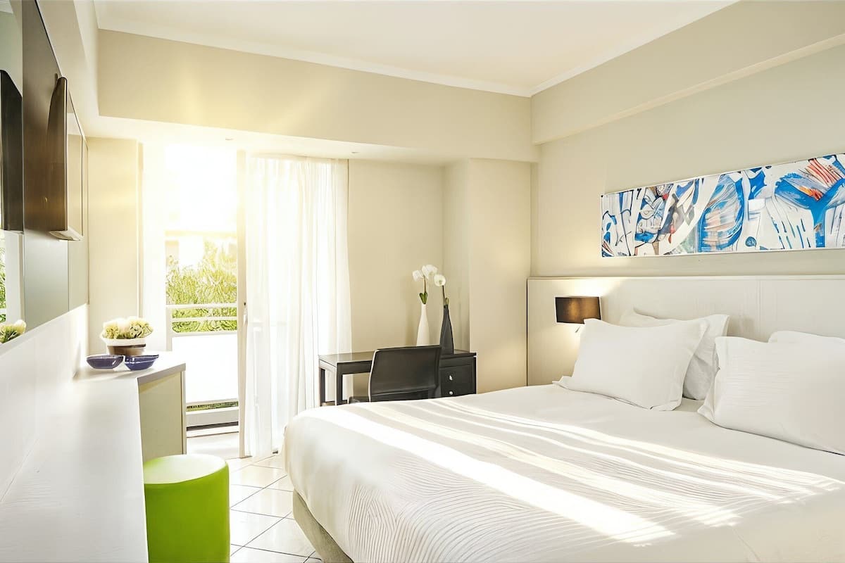 Ein Hotelzimmer mit Bett, Schreibtisch, Stuhl, einem kleinen Tisch mit Vase und einem Bild an der Wand.