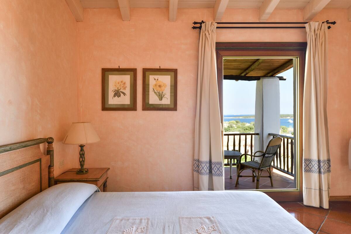 A bedroom with a bed, a nightstand with a lamp, two framed floral pictures on the wall, and a balcony with a chair.