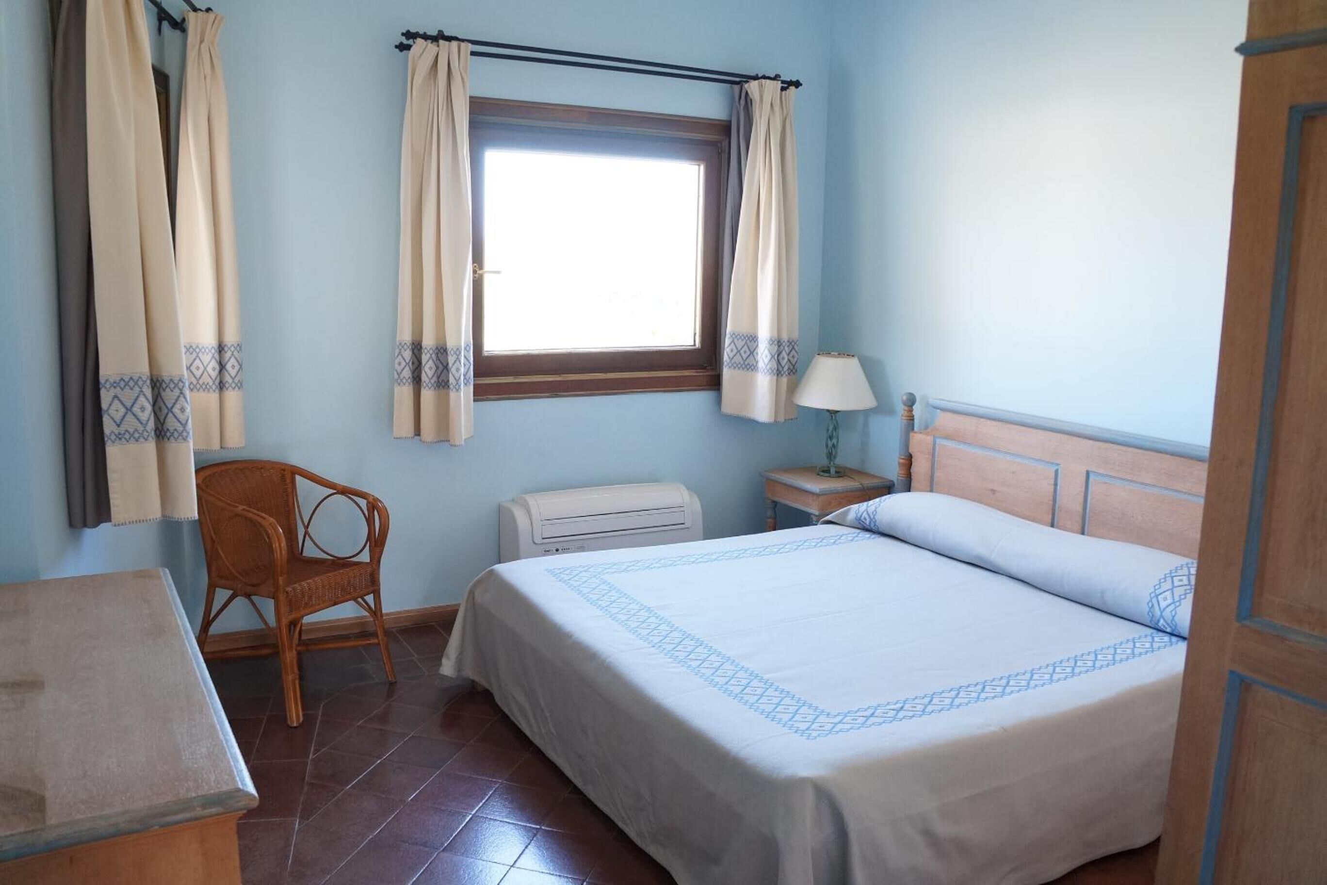 A room with a bed, a chair, a table, a window with curtains, and an air conditioning unit.