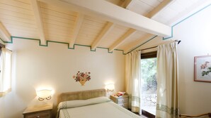 A bedroom with a bed, bedside tables, a nightstand, a wall-mounted lamp, a window with curtains, and a framed picture on the wall.