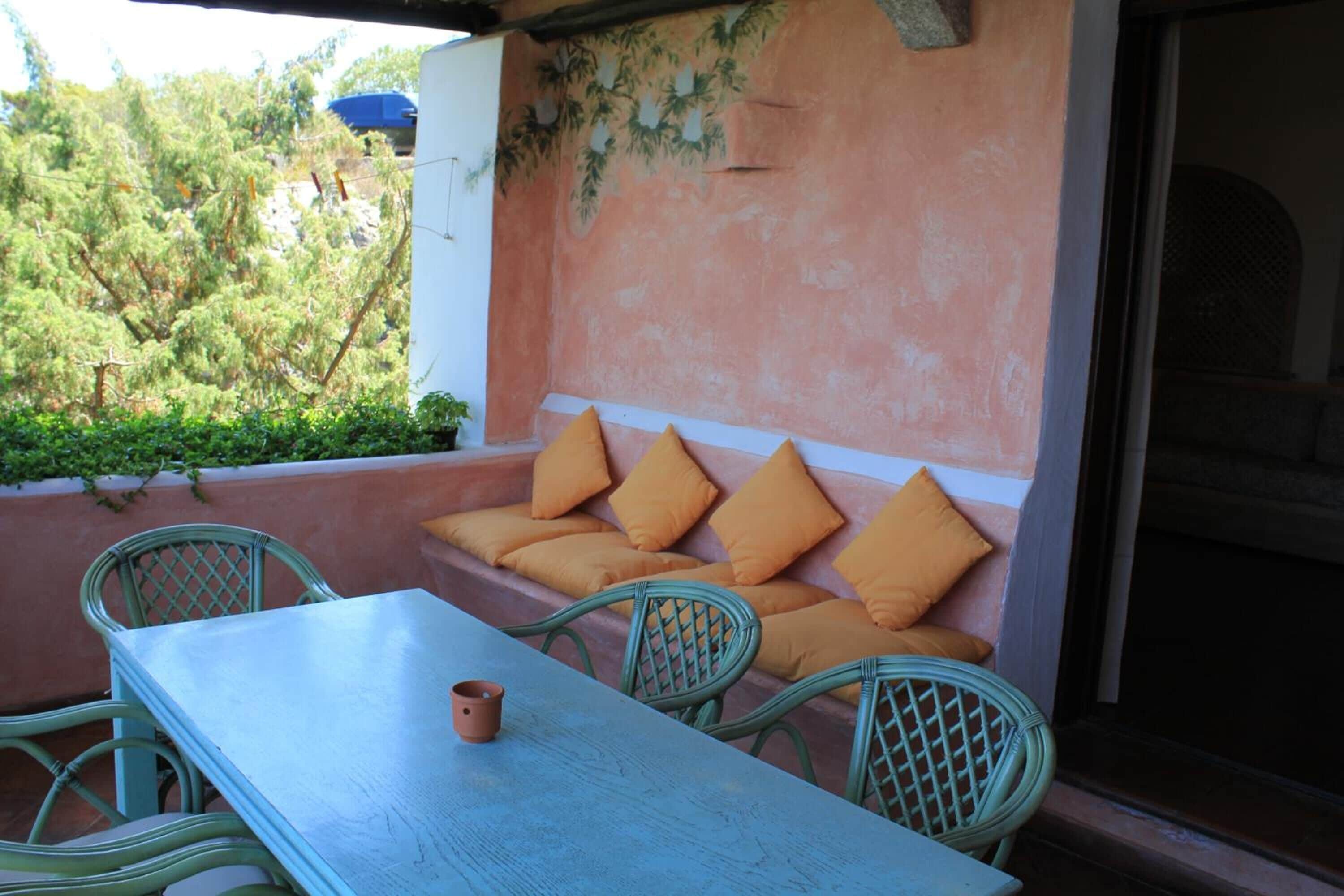 Un área de estar al aire libre con una mesa y sillas, un banco acolchado con cojines anaranjados y una pared con un mural.