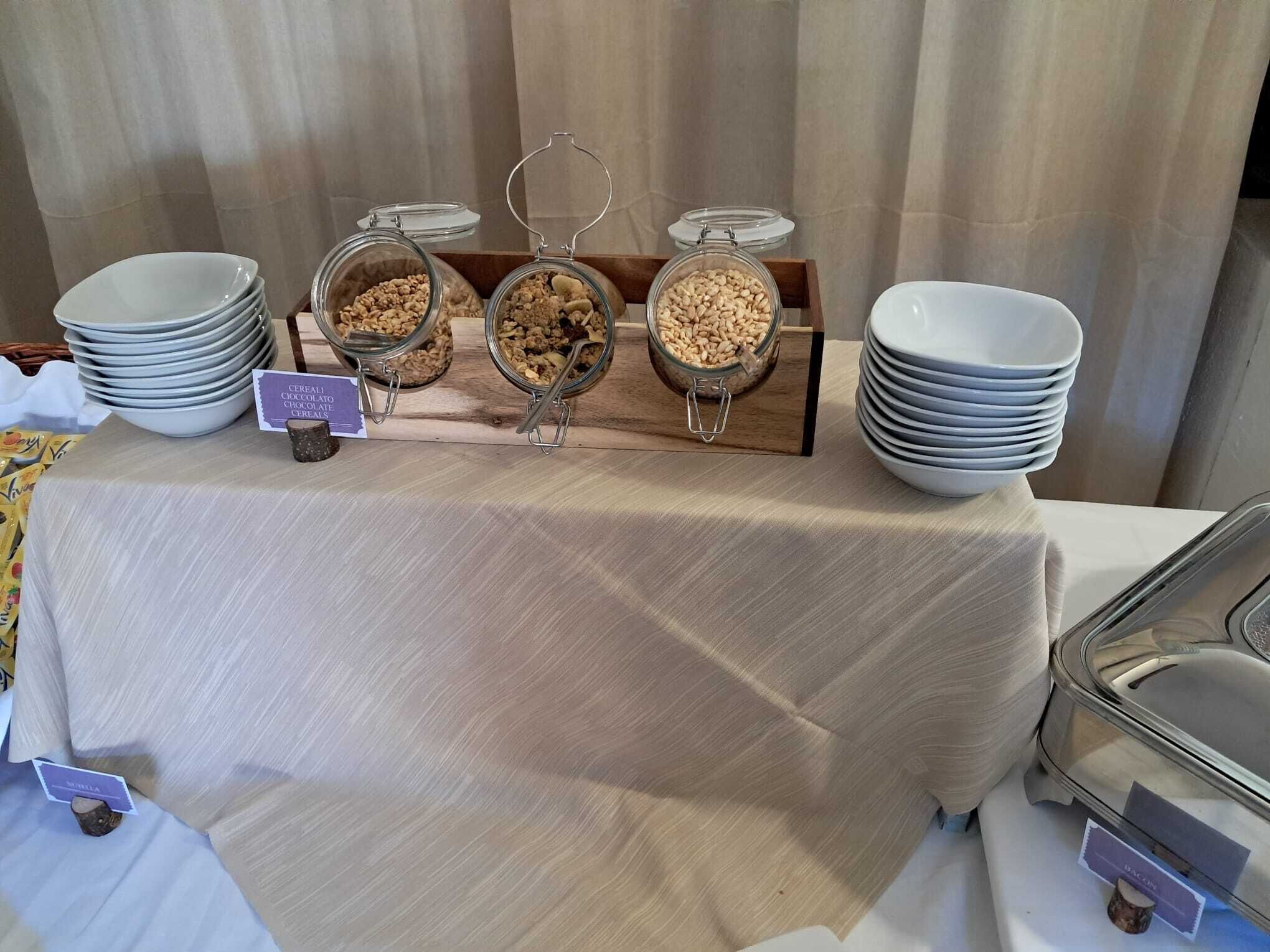A buffet table with three glass containers labeled with different types of cereal and a stack of white plates on either side.