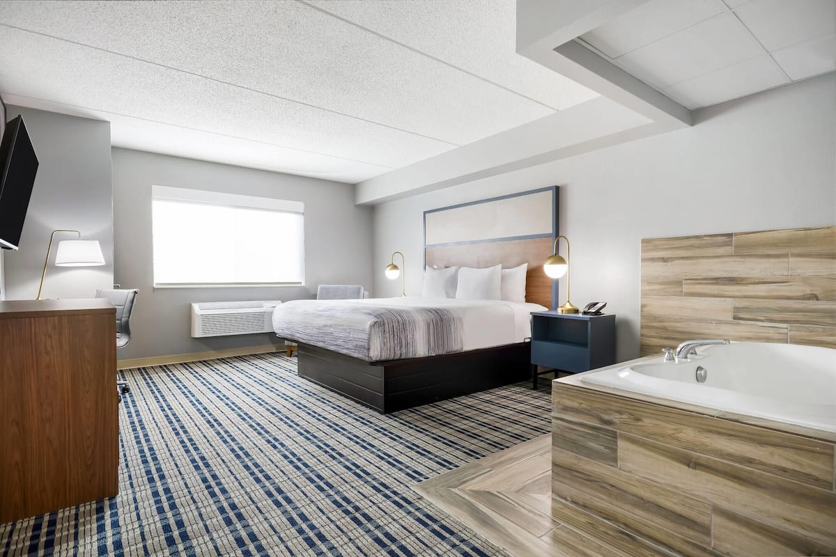 Een moderne hotelkamer met een groot bed, een inbouw bureau, een flatscreen televisie, een groot raam, een badkamer met bad en een wandpaneel van hout.