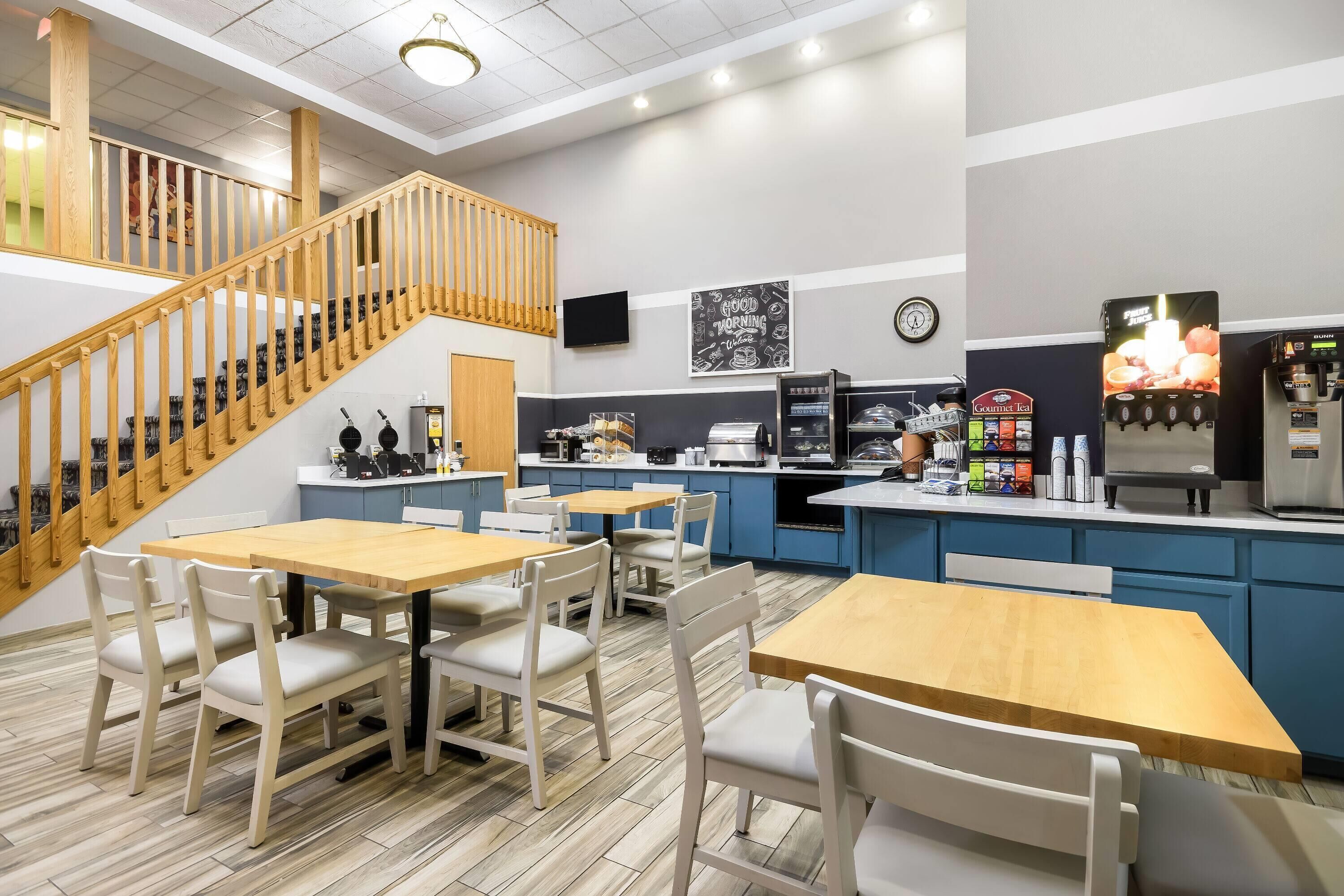 Un café con mesas y sillas de madera, una escalera, una zona de cocina y un dispensador de zumo de frutas.