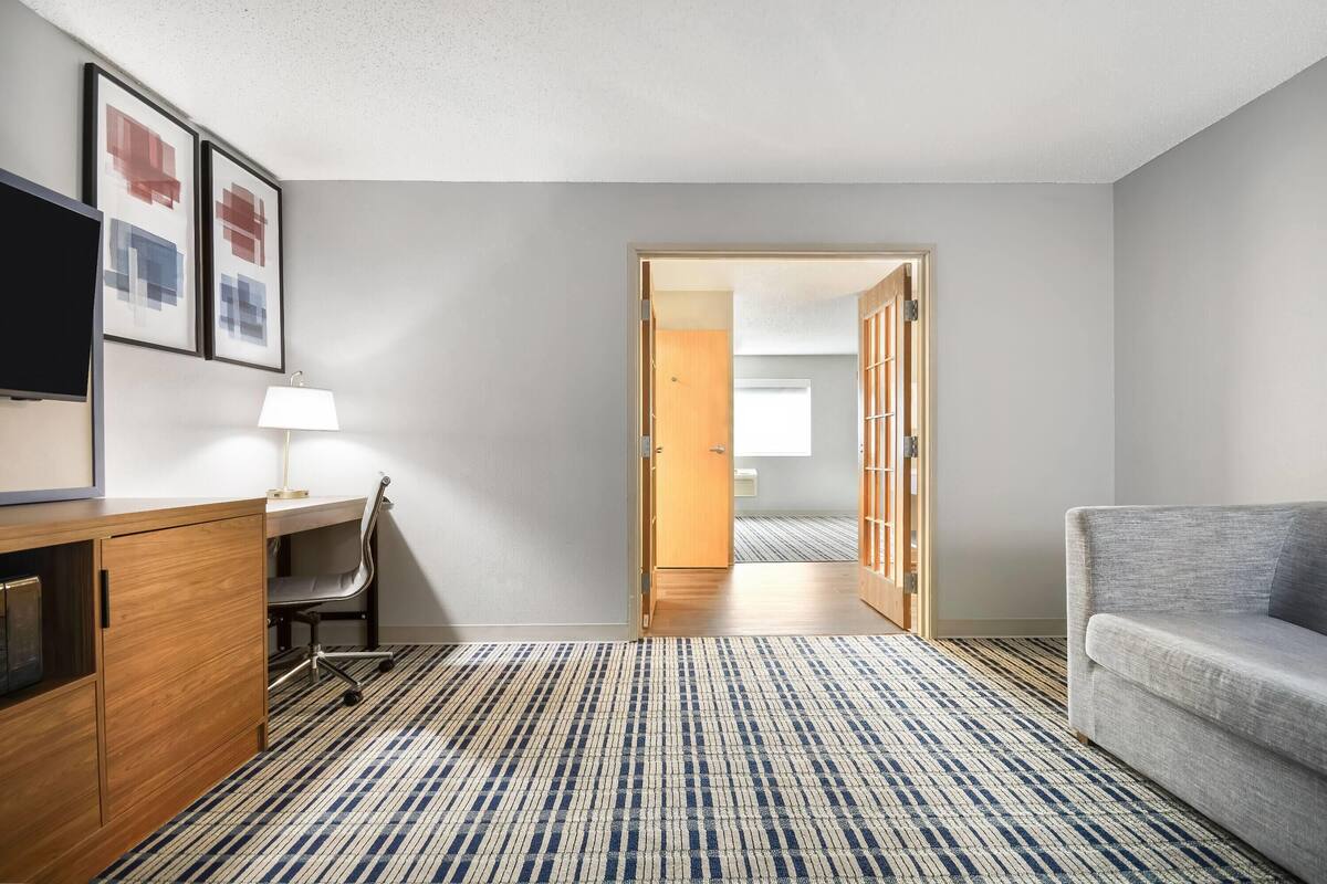 Een hotelkamer met een bureau, stoel en een bank. Er is een deur die naar een andere kamer leidt.