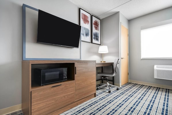 Une chambre d’hôtel équipée d’une télévision à écran plat, d’un four à micro-ondes, d’un bureau avec une chaise, d’une lampe et d’une fenêtre avec un climatiseur.