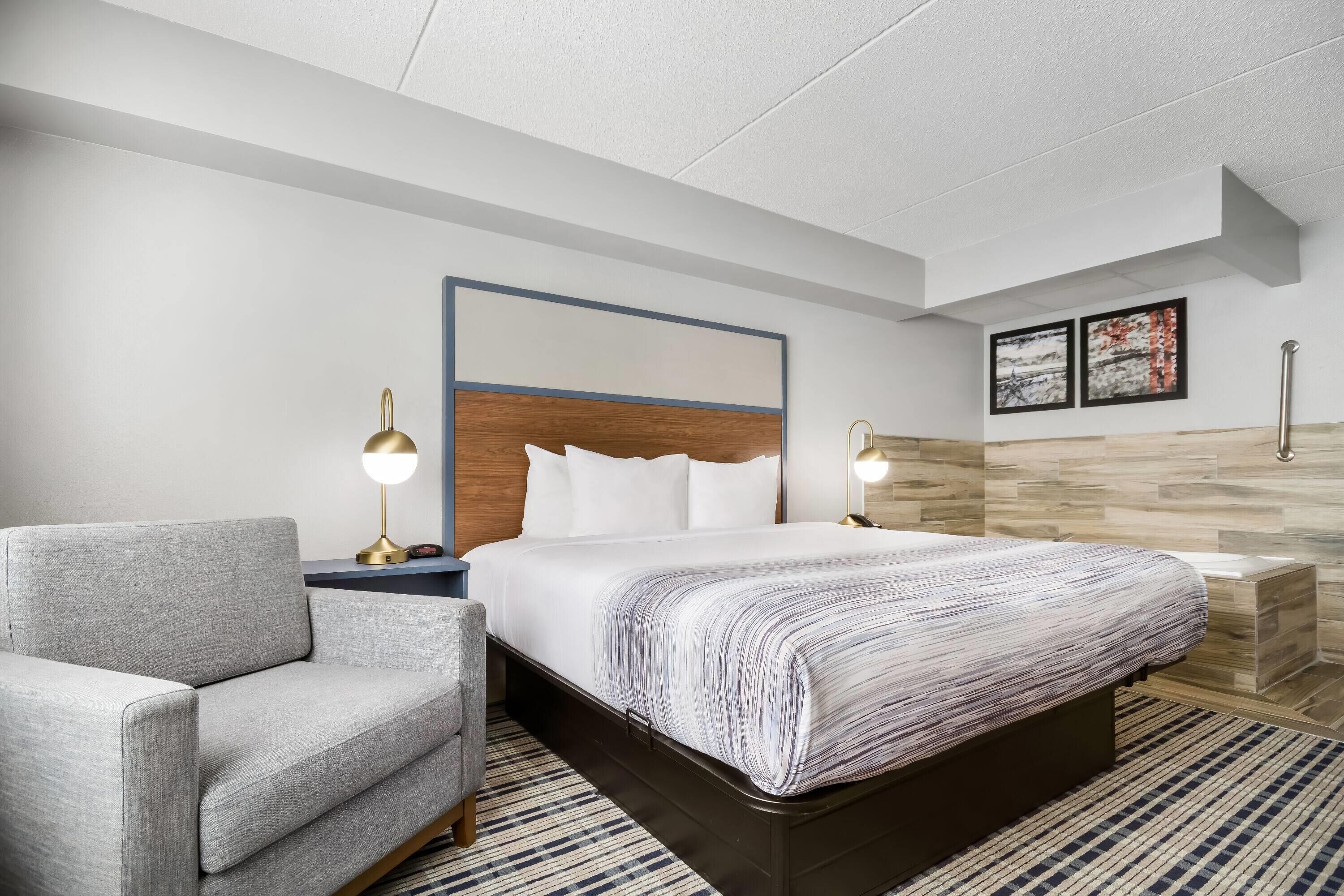 Quarto de hotel moderno com uma cama grande, uma poltrona cinza, uma mesa de cabeceira com abajur e uma cabeceira de madeira.