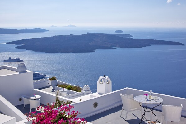 A rooftop terrace with white furniture, a table with a vase, and a view of the sea and islands.