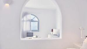 A minimalist room with a large arched window, a bed, a bedside table, and a lamp. - Cliff Side Suites (Santorini)
