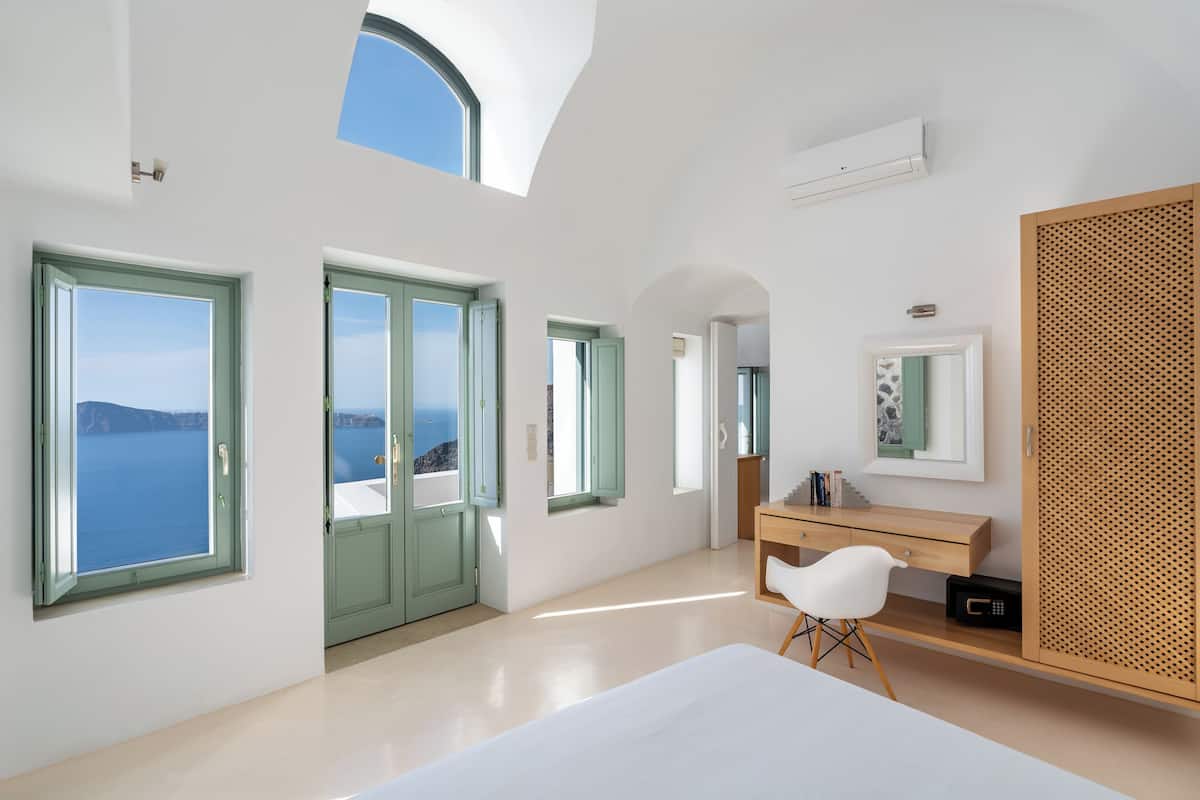 A modern hotel room with large windows, a wooden desk, a white chair, and a bed.