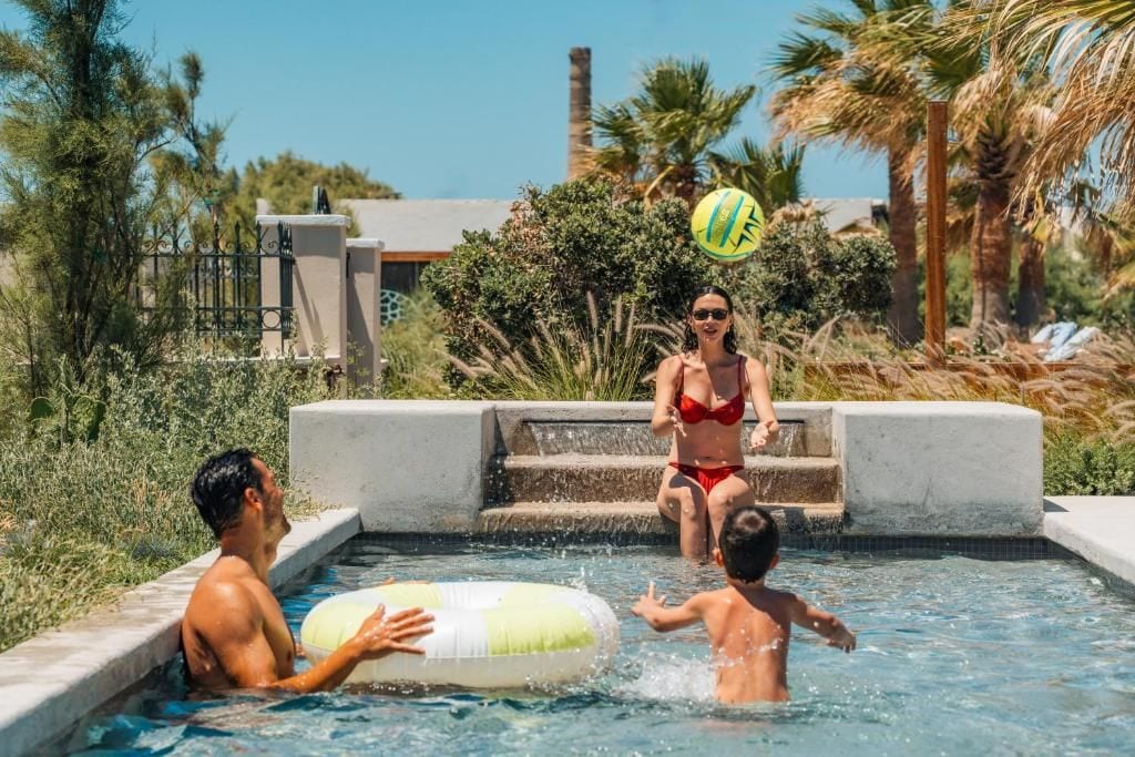 A swimming pool with a person on a floating platform, another person standing on the edge, and a third person in the water.