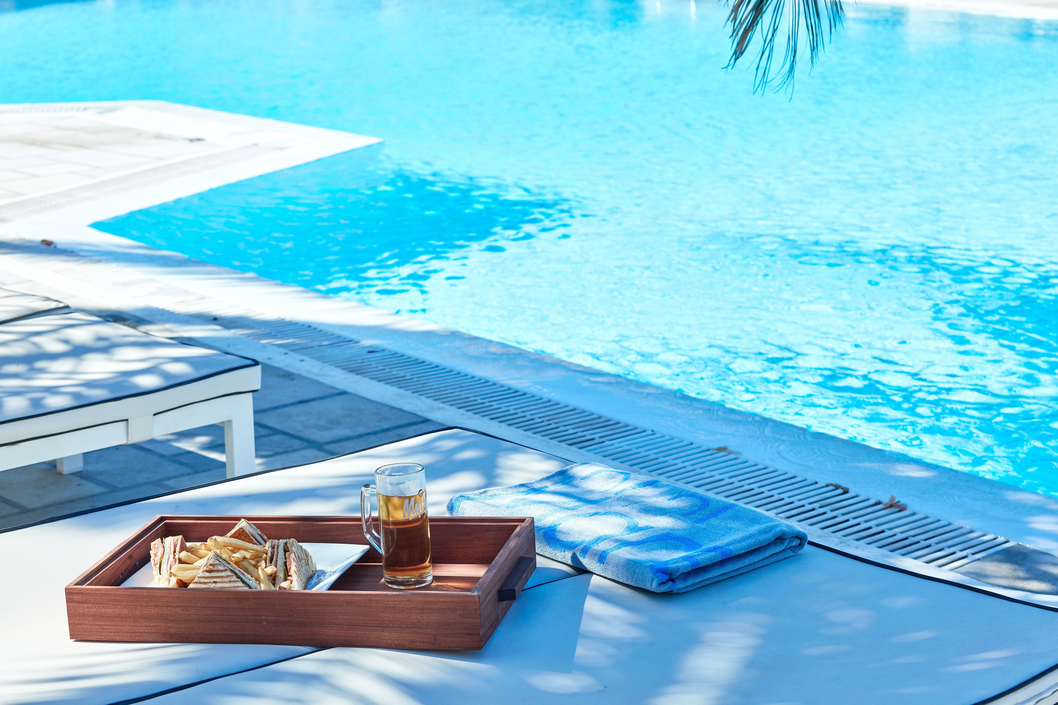 A poolside setting with a wooden tray containing sandwiches and a glass of beer, a towel, and a lounge chair.