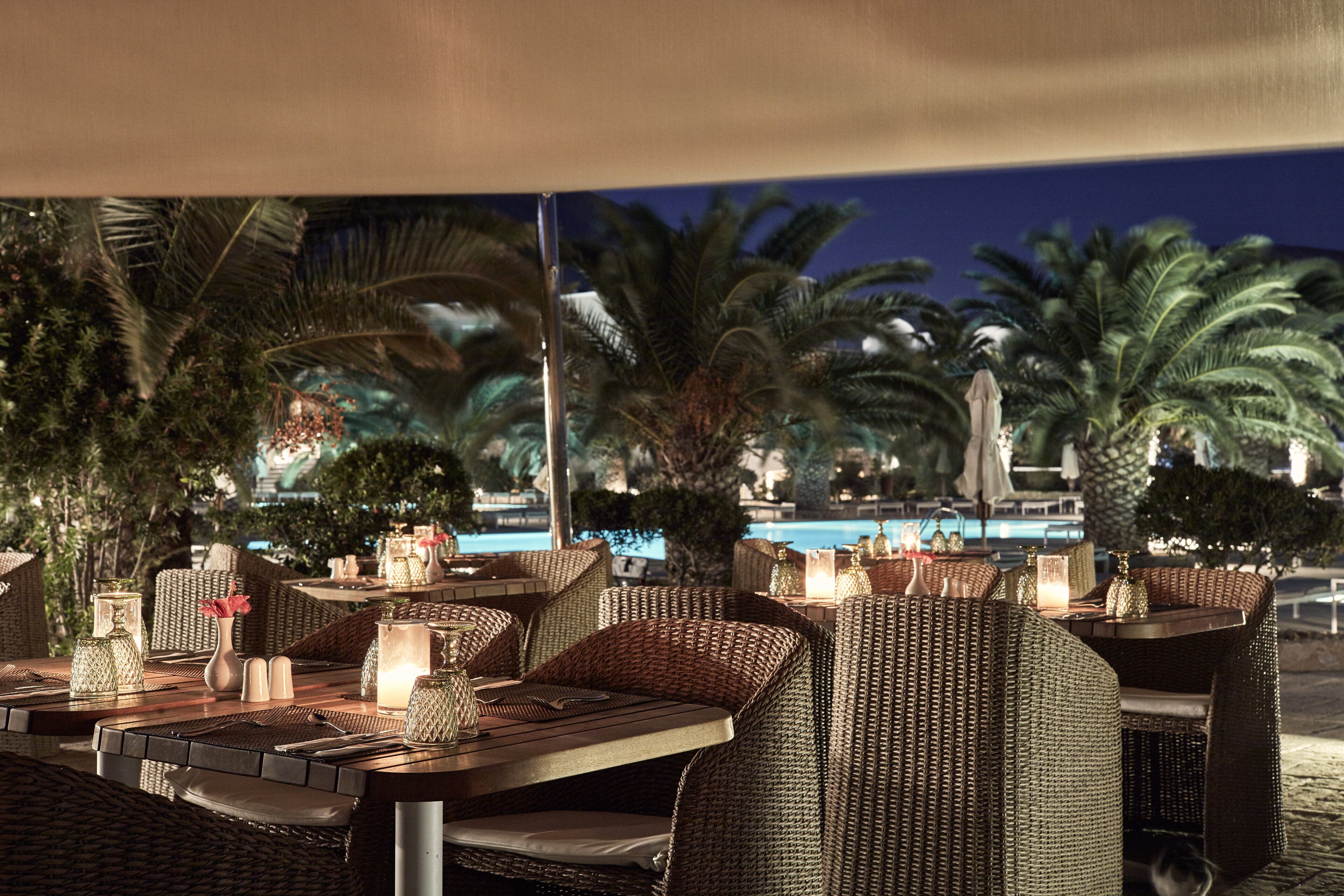 An outdoor dining area with wicker furniture, candles, and a view of a pool at night.