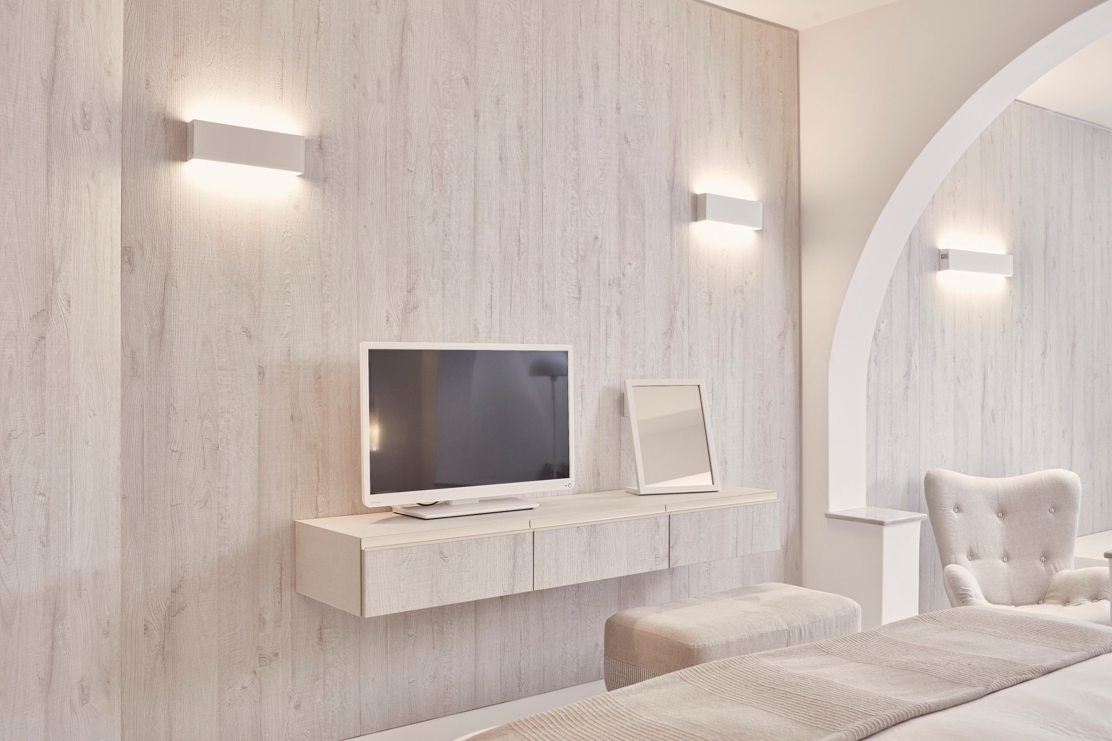 A modern bedroom with a TV on a floating shelf, a bedside table, and a tufted armchair.