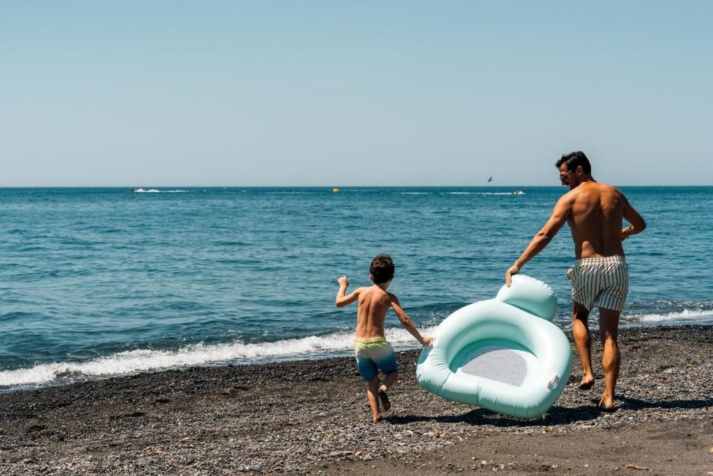 Escena de playa con un niño y un adulto caminando hacia el agua, llevando un flotador.