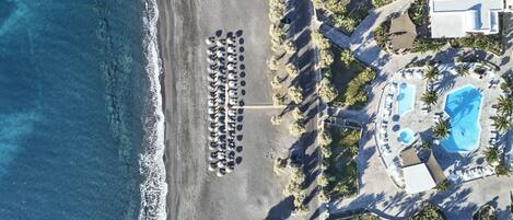 An aerial view of a coastal hotel with a beach, a swimming pool, and a parking area.