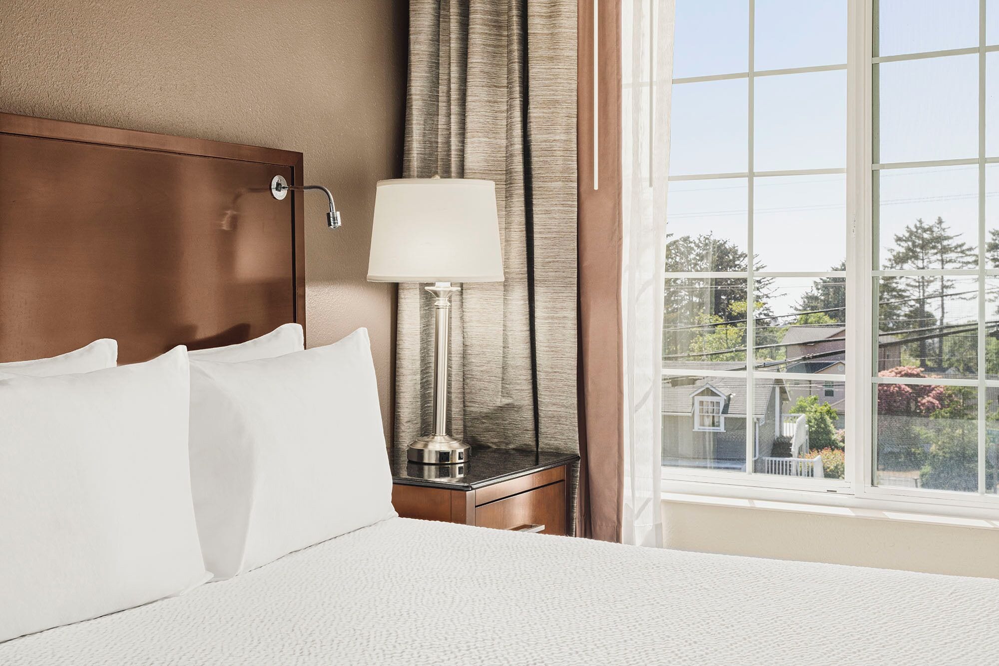 A hotel room with a bed, a nightstand, a lamp, and a window with a view of buildings and trees.