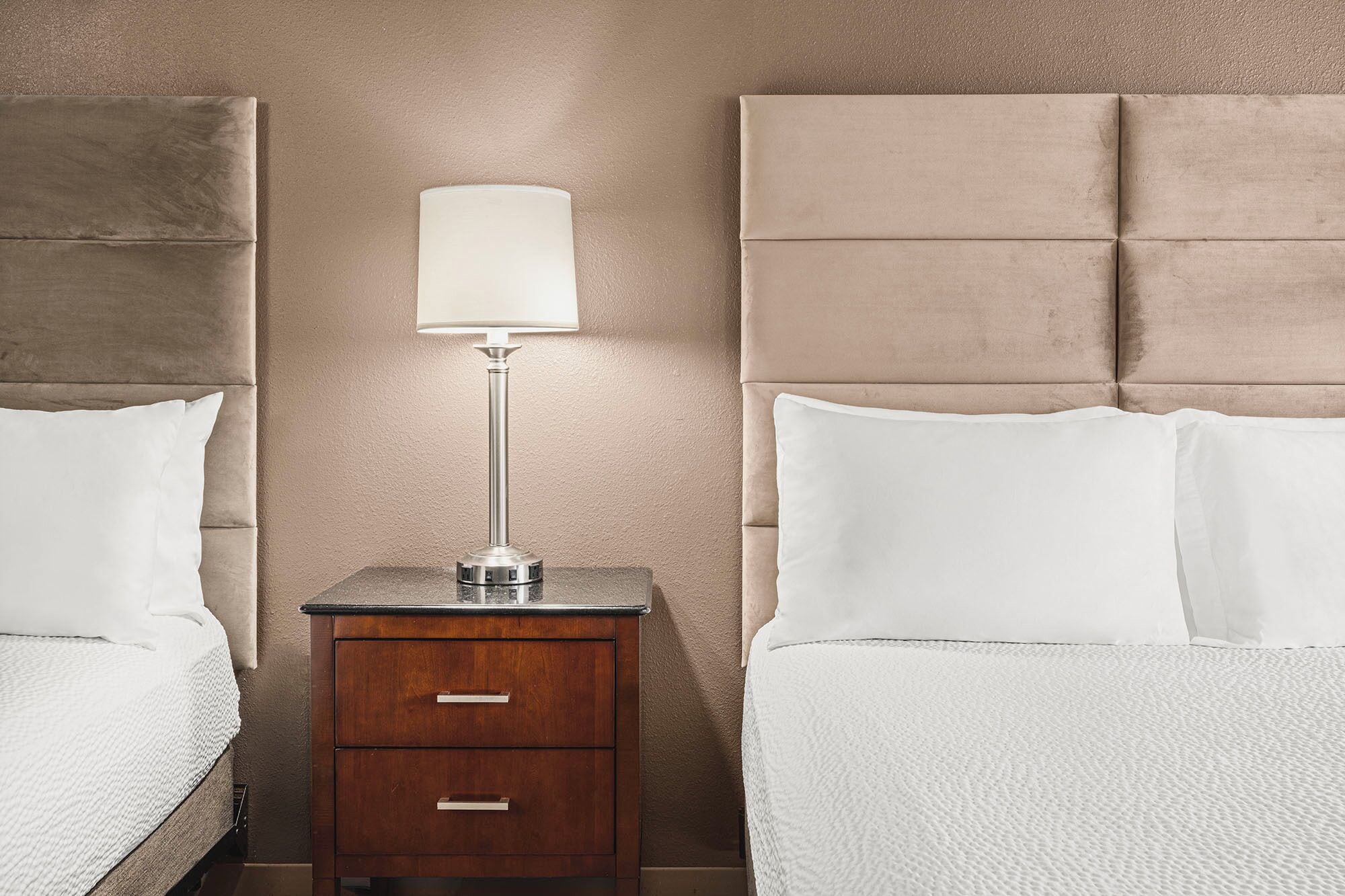 Quarto de hotel com duas camas, mesa de cabeceira com abajur e cômoda de madeira.