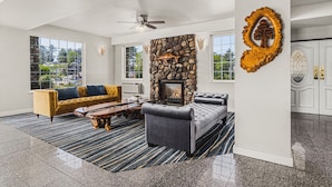 A spacious living room with a stone fireplace, a ceiling fan, and a large window offering a view of the outdoors. - The Ashley Inn & Suites (Lincoln City)