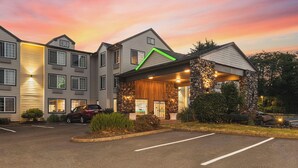 A multi-story hotel with a stone facade entrance, a parking lot, and a sunset sky. - The Ashley Inn & Suites (Lincoln City)