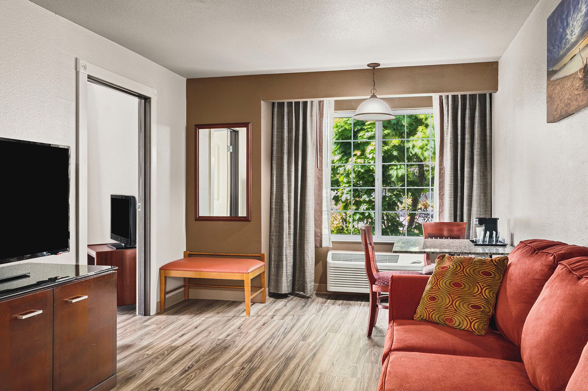 Een hotelkamer met een houten vloer, een rode bank, een eettafel met stoelen, een televisie, een raam met gordijnen en een plafondlamp.