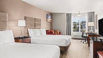Ett hotellrum med två sängar, ett skrivbord, en stol, en TV och ett fönster med utsikt över byggnader och träd.