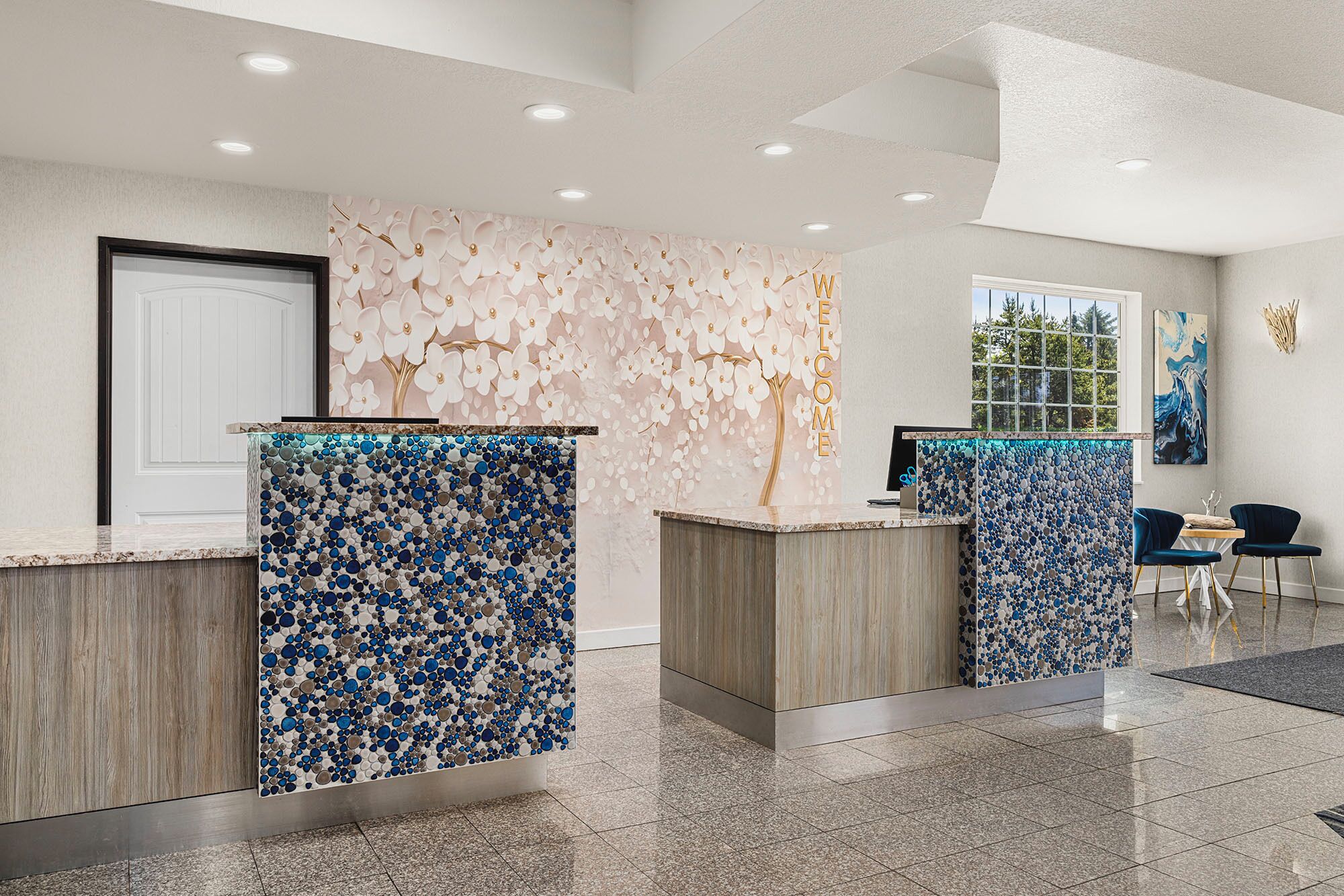 A reception area with two counters, one with a blue and white mosaic design and the other with a wooden finish. There is a door to the left and a window with a view of trees to the right.