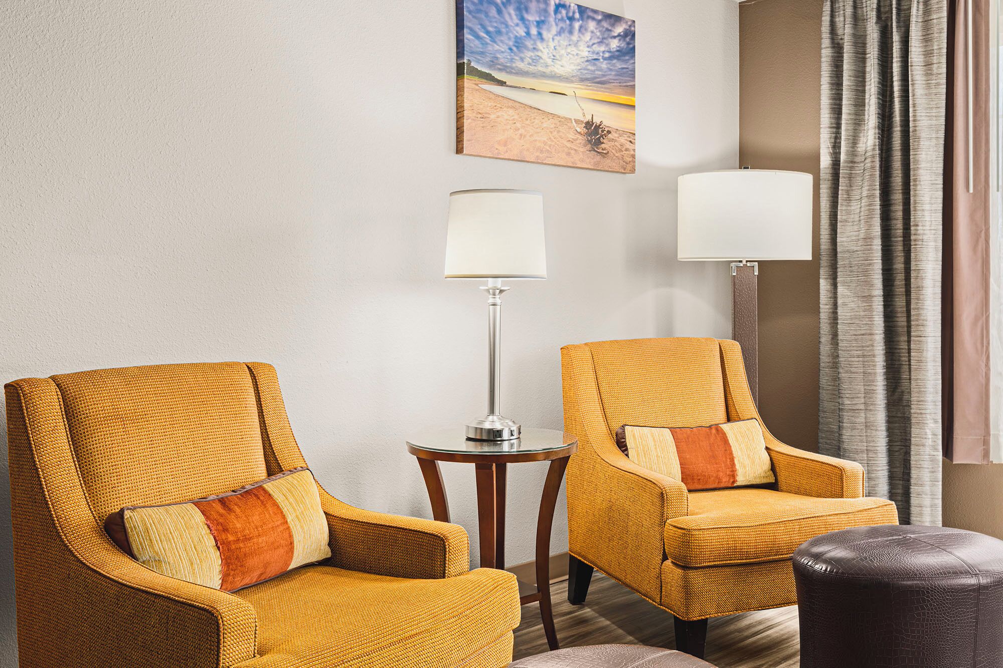 A room with two yellow armchairs, a small table, two lamps, and a beach painting on the wall.