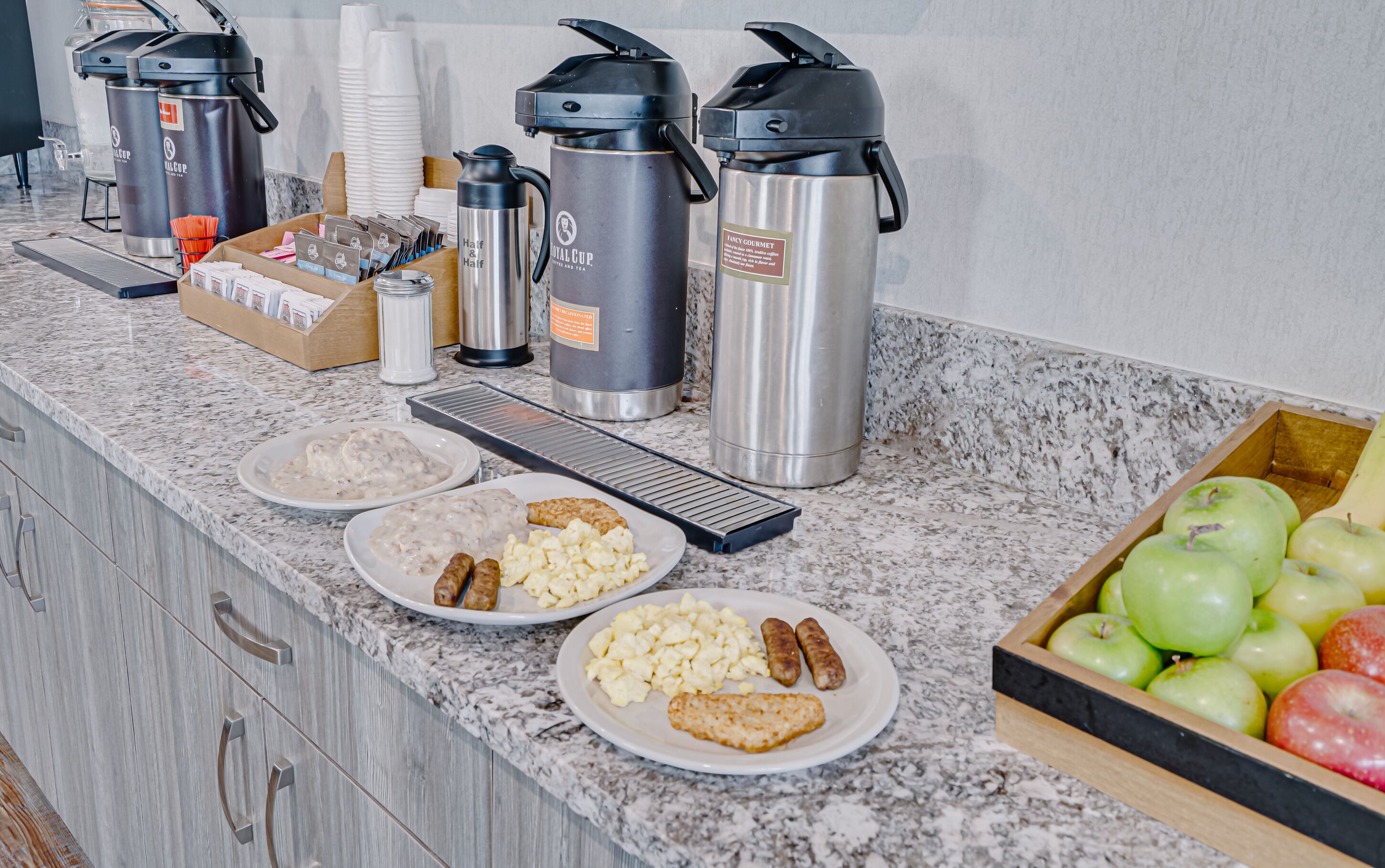 A breakfast buffet setup with various food items, coffee makers, and a fruit basket.