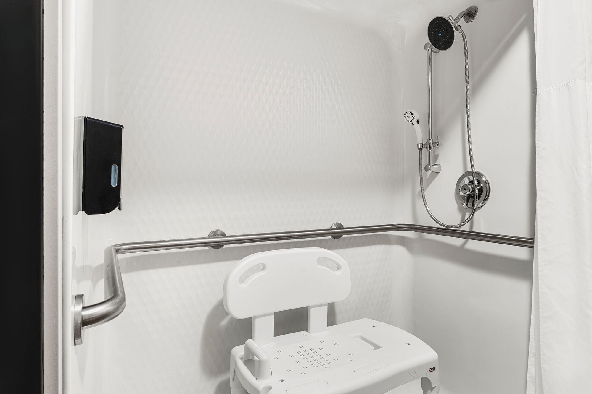 A bathroom with a white shower enclosure, a handheld showerhead, and a white toilet with a safety rail.