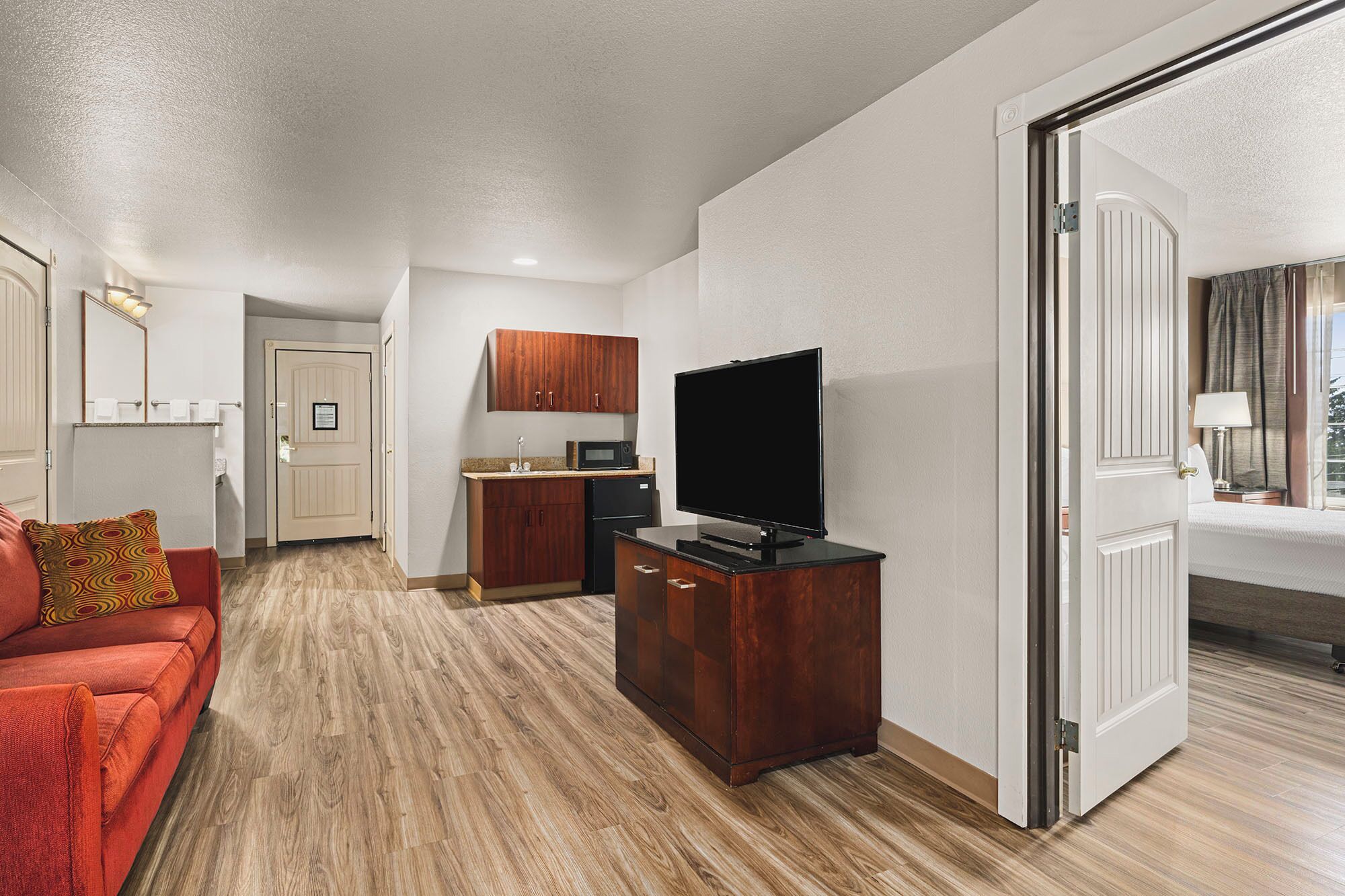 A compact living space with a kitchenette, a flat-screen TV, a red sofa, and a wooden floor.