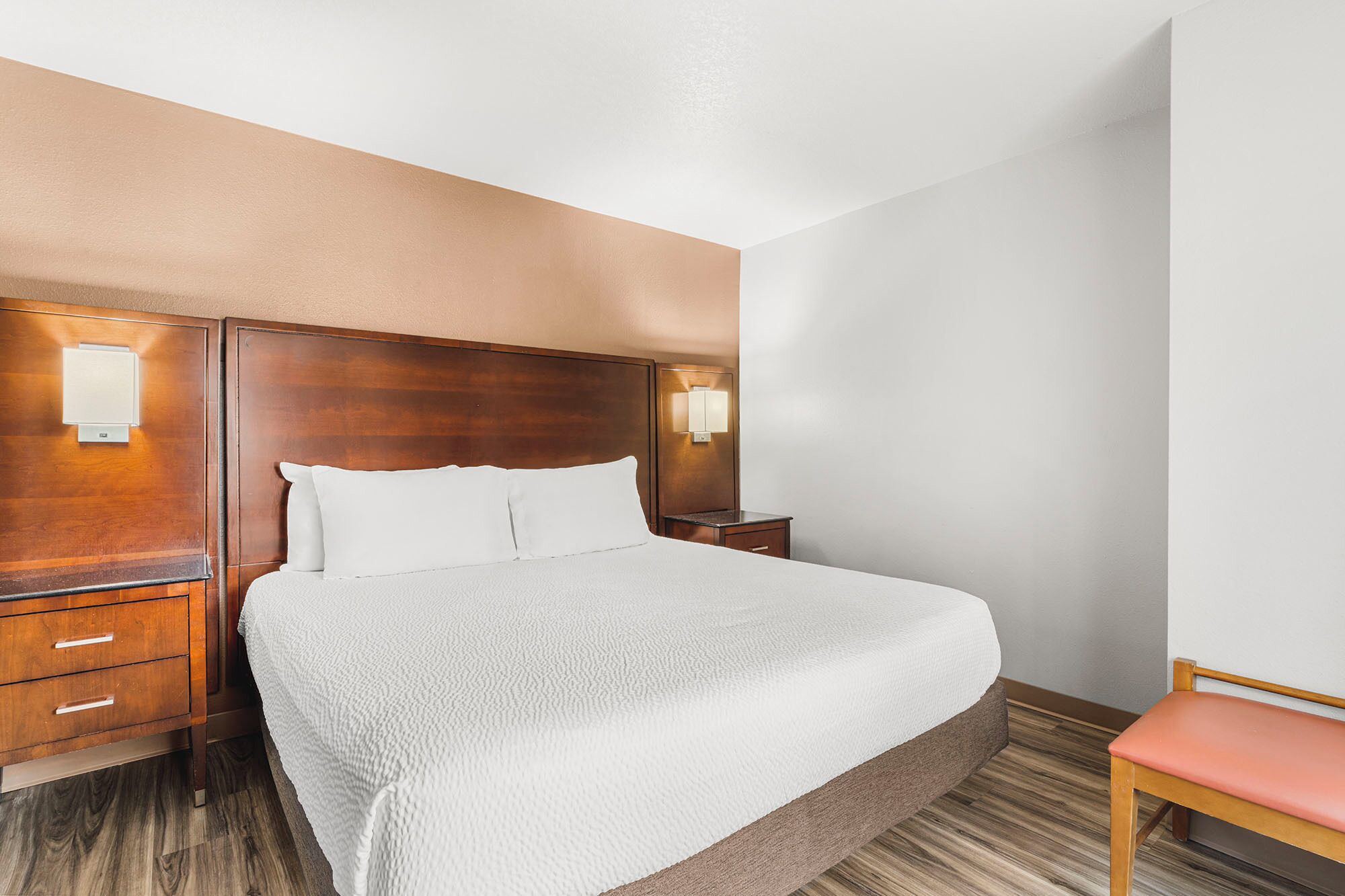 A hotel room with a large bed, wooden headboard, two bedside tables with lamps, a wooden nightstand, and a wooden chair.