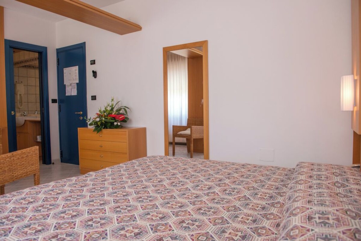 Une chambre avec une couvre-lit à motifs, une commode en bois ornée d’une plante, une porte bleue et un miroir.