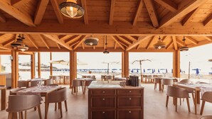 Un vaste espace de restauration intérieur, agrémenté de poutres en bois et de grandes fenêtres donnant sur une plage. Les tables sont dressées avec des nappes blanches et des chaises.
