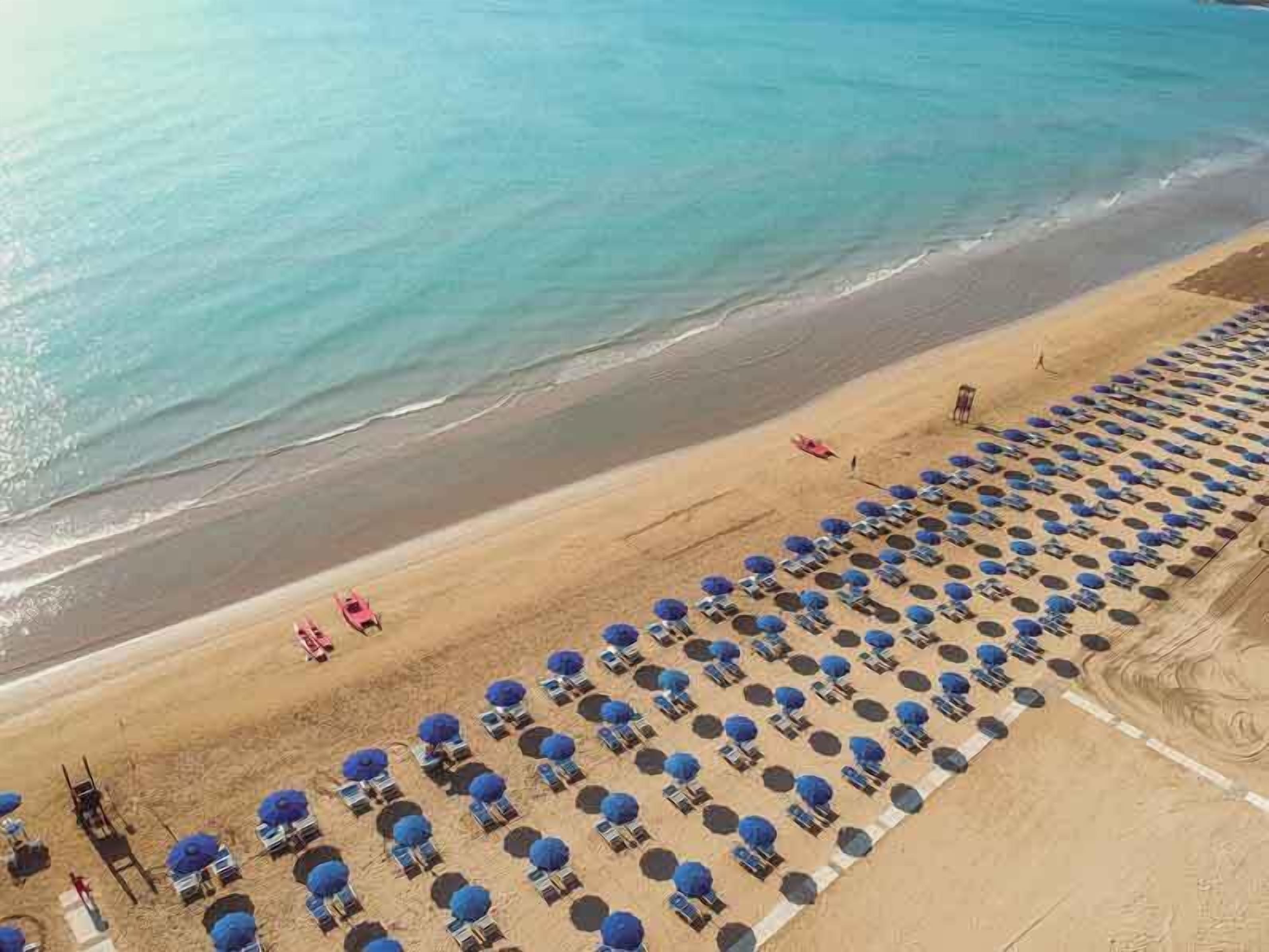 Vista aérea de uma praia com fileiras de guarda-sóis azuis e espreguiçadeiras, uma faixa de areia e o oceano.