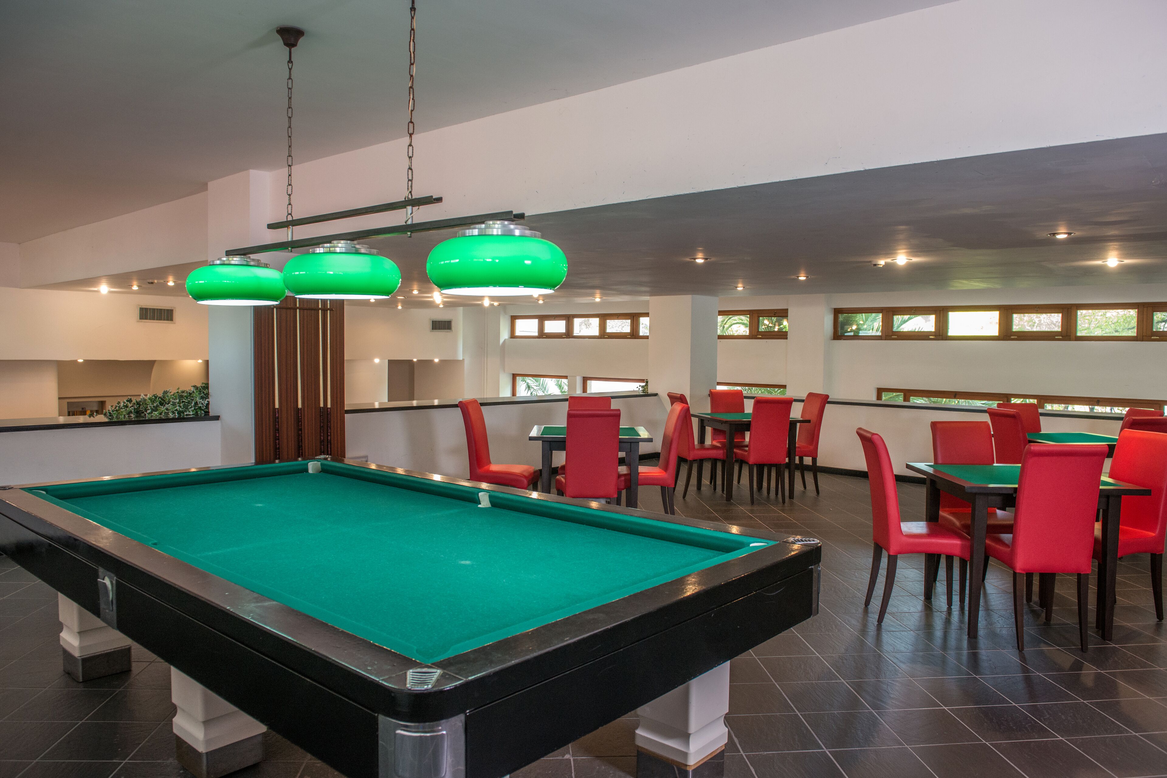 A pool table in a room with red chairs and green tables, with a ceiling light fixture above.