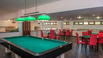 A pool table in a room with red chairs and green tables, with a ceiling light fixture above.