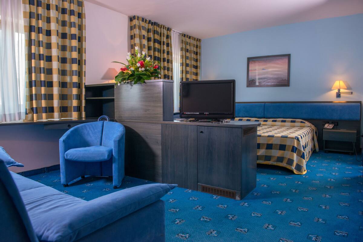 Ein Hotelzimmer mit einem blauen Sofa, einem blauen Sessel, einem Bett mit karierter Bettdecke, einem Fernseher auf einem Holzschrank, einer Vase mit Blumen und einem Gemälde an der Wand.