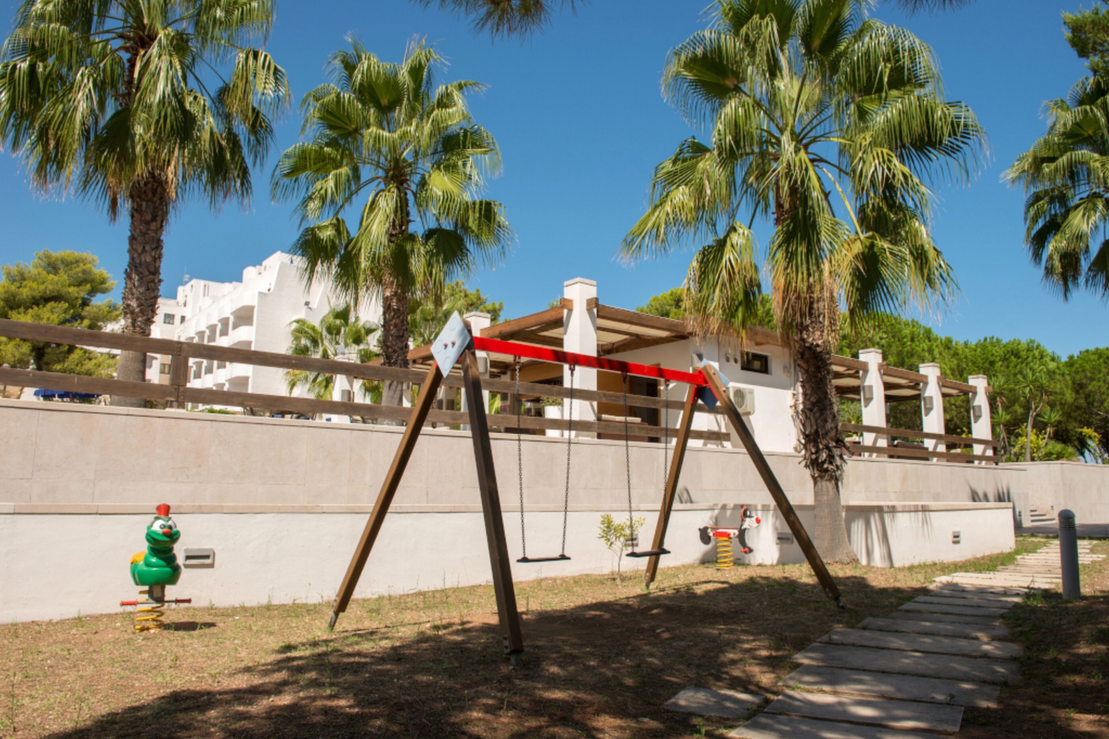 A playground with swings and a slide, surrounded by palm trees and a white building in the background.