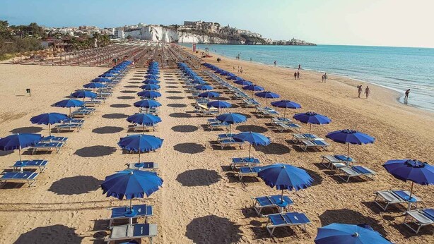Une plage bordée de rangées de transats et de parasols, un ciel dégagé et des bâtiments en arrière-plan.