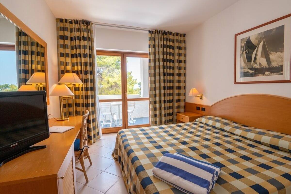 Ein Hotelzimmer mit kariertem Bette, einem Fernseher auf einem Holztisch, einer Schreibtischlampe, einem Stuhl und einem Balkon mit Vorhängen.